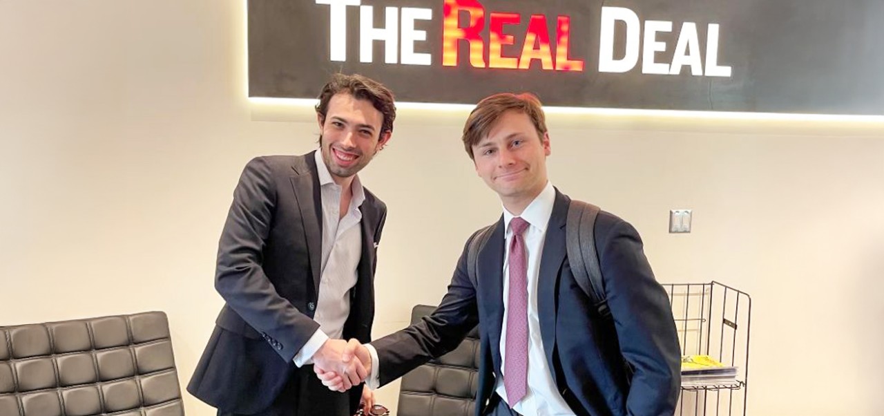 Harrison Koeppel, Schack Institute at NYU SPS alumnus, shakes hands with a colleague in front of a logo for the Real Deal publicaion.
