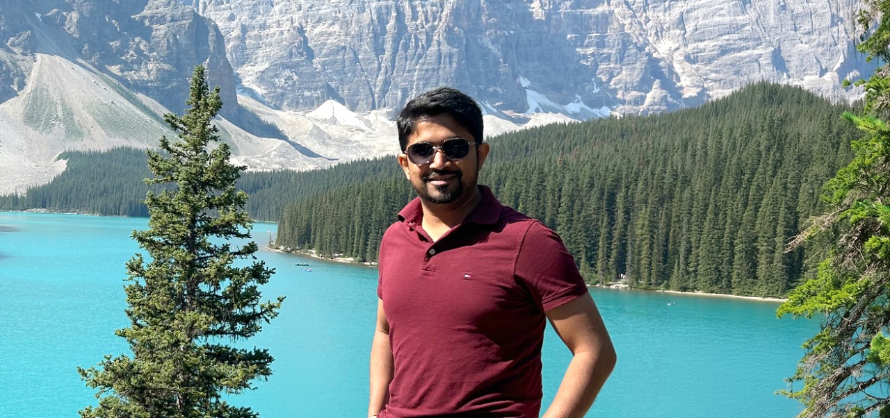 NYU SPS Division of Programs in Business alumnus Samarth Bedakihale poses for a photo in front of a scenic lake surrounded by trees and mountains.