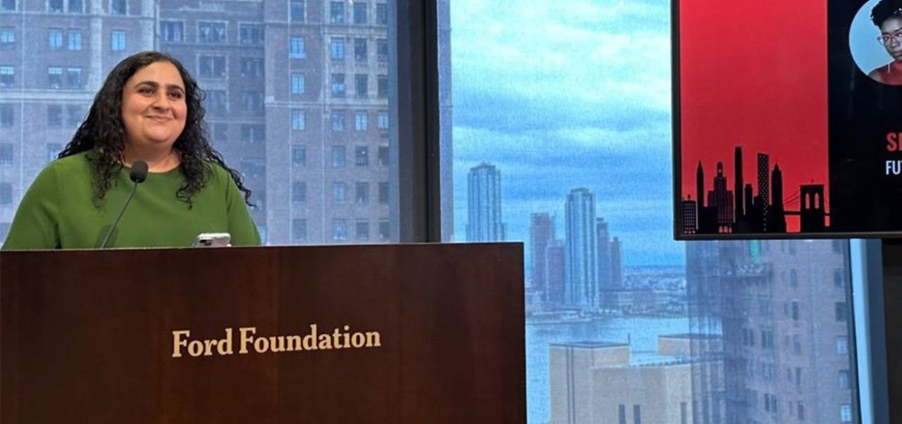 Sandra Khalil, Center for Global Affairs at NYU SPS alumnus, stands at a lecturn at the Ford Foundation in front of a glass window revealing midtown Manhattan skyscrapers.
