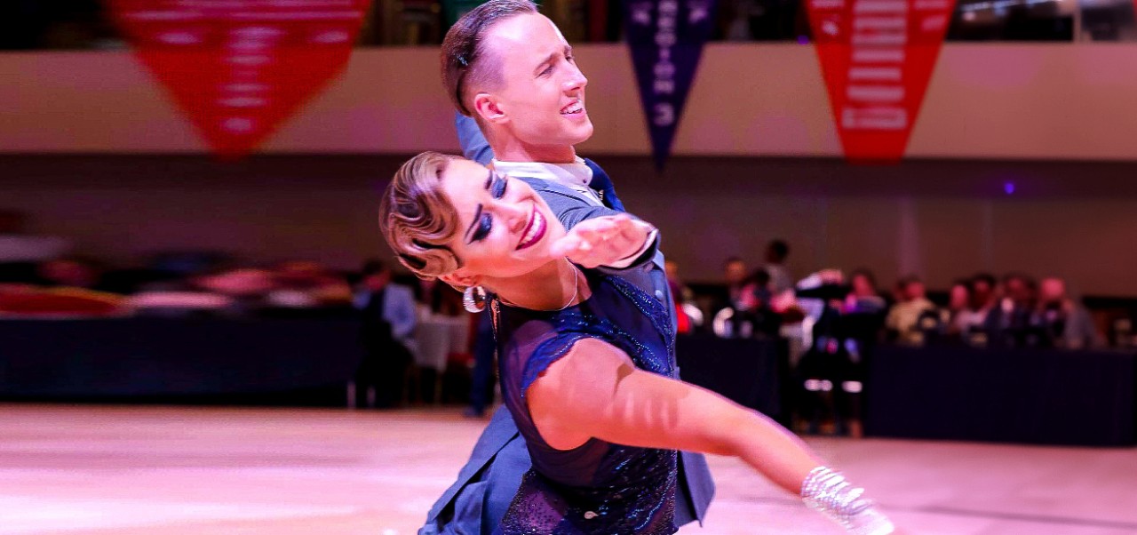 Talia Barrington, NYU SPS Division of Applied Undergraduate Studies alumnus, dances with a male partner on a stage inf a crowded ballroom.