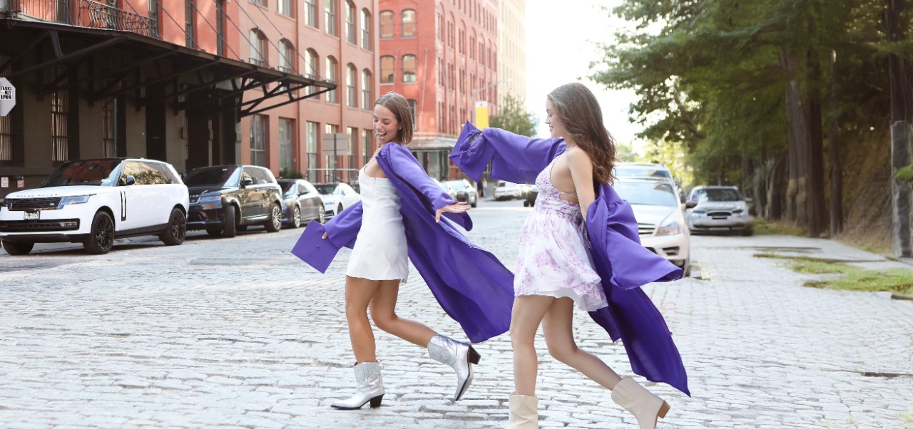 Jonathan Tisch Center of Hospitality alumnus Yasmine Barbes and a friend stroll through a cobblestone New York City street while wearing graduation regalia.