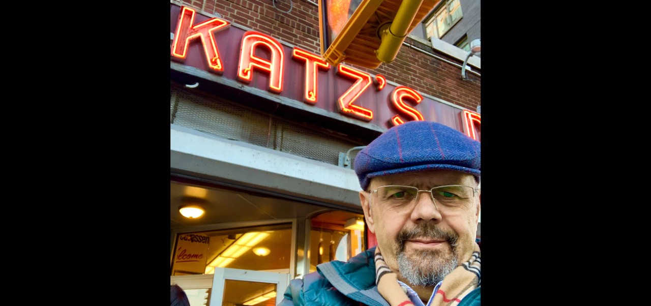 Andres Fortino takes a selfie wearing a blue hat, scarf, and coat in front of a red neon sign that reads "Katz's"