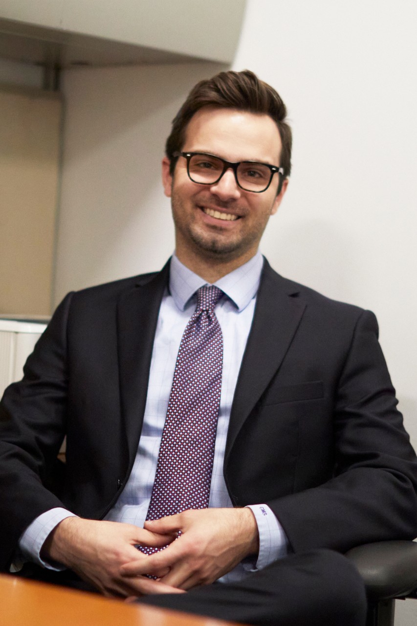 Colin Slabach sits with his hands folded in front of him for a headshot in an office setting