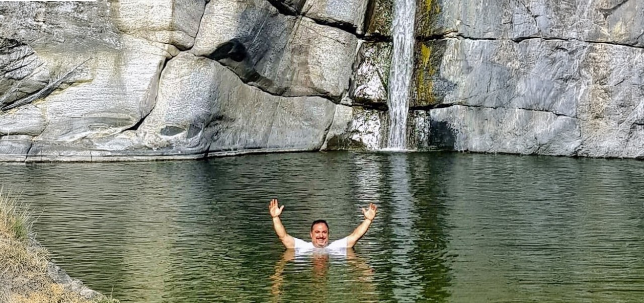 While swimming in a pond with a small waterfall and a large cliff behind him, Dave Lipsky smiles with his hands above his head