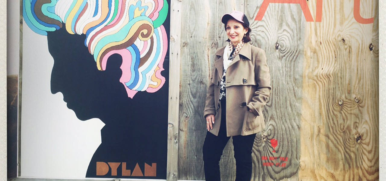 Mary Cianni wears a trench coat, black pants, and a baseball cap while standing next to a colorful mural of Bob Dylan at an art museum