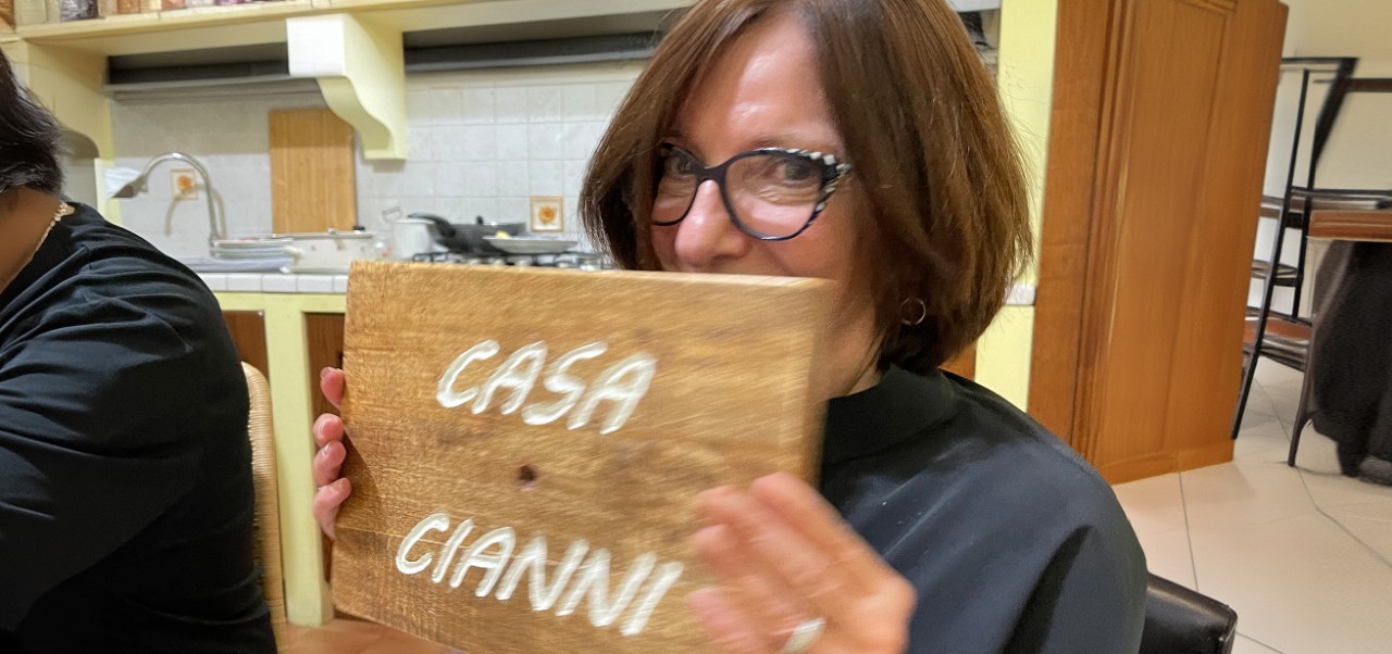 Mary Cianni covers half of her face with a wooden board that says "Casa Cianni" while visiting the hometown of her grandfather in Italy