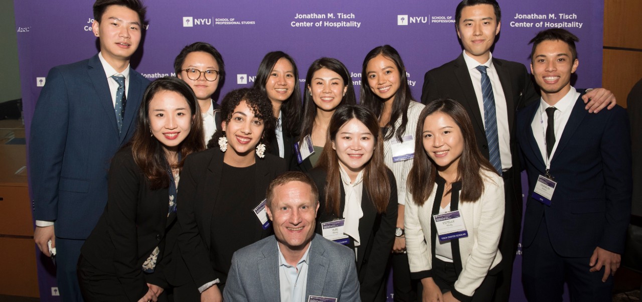 Nicolas Graf, Clinical Professor, Jonathan M. Tisch Center of Hospitality at NYU SPS poses for a photo with ten students in front of a purple step and repeat.