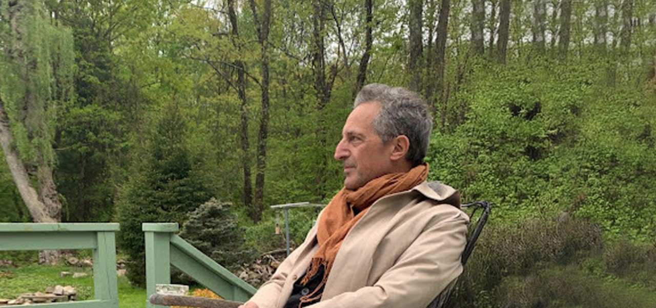 Sitting on a porch with a forest behind it, Pierre Lacour wears a beige jacket and orange scarf while looking to the left side of the image.