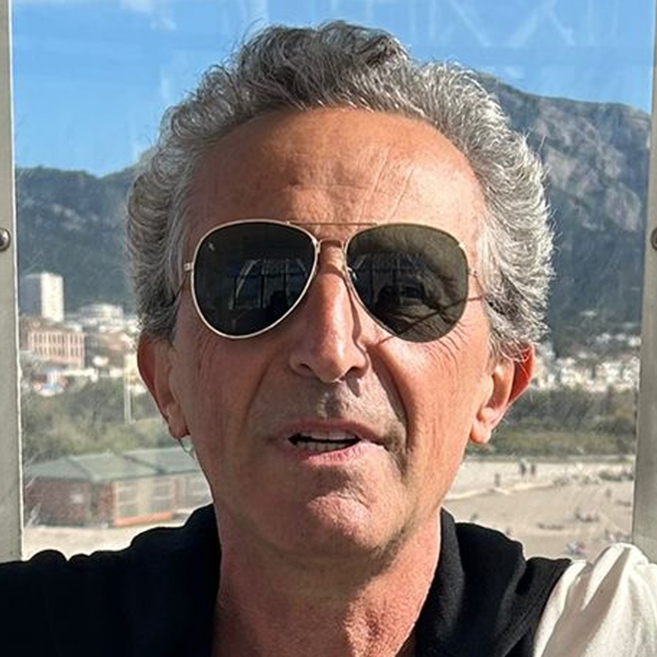 Standing in front of a window with a mountainous scene behind him, Pierre Lacour slightly smiles for a photo while wearing a white t-shirt with a black sweater tied around his shoulders and sunglasses