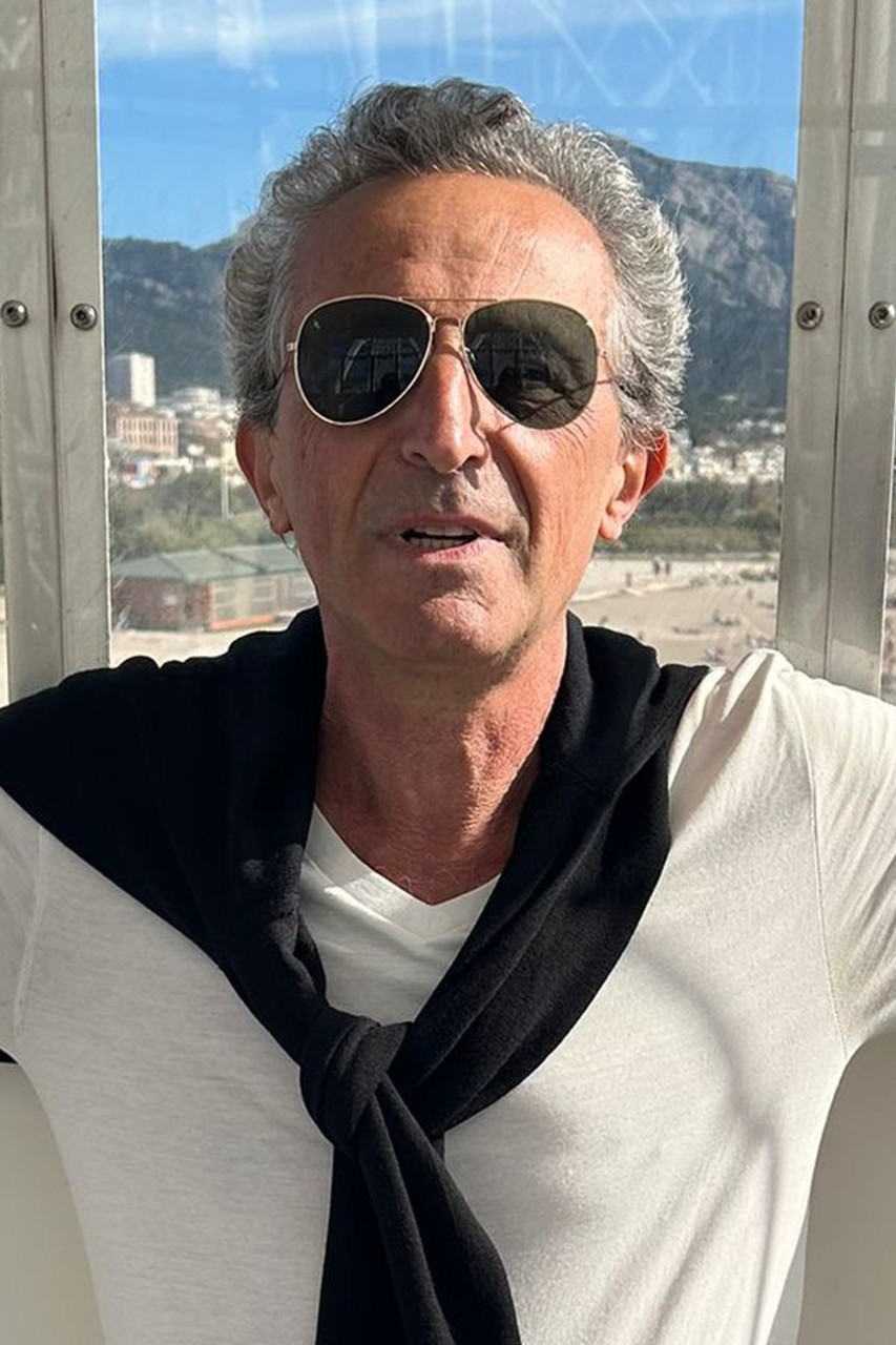 Standing in front of a window with a mountainous scene behind him, Pierre Lacour slightly smiles for a photo while wearing a white t-shirt with a black sweater tied around his shoulders and sunglasses