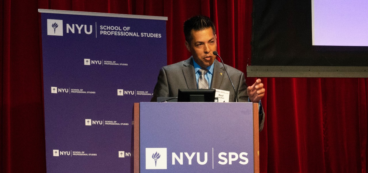 Raul Sanchez speaks into a microphone onstage behind a purple podium with a red curtain and an NYU SPS banner behind him at the NYU SPS Innovation Day event