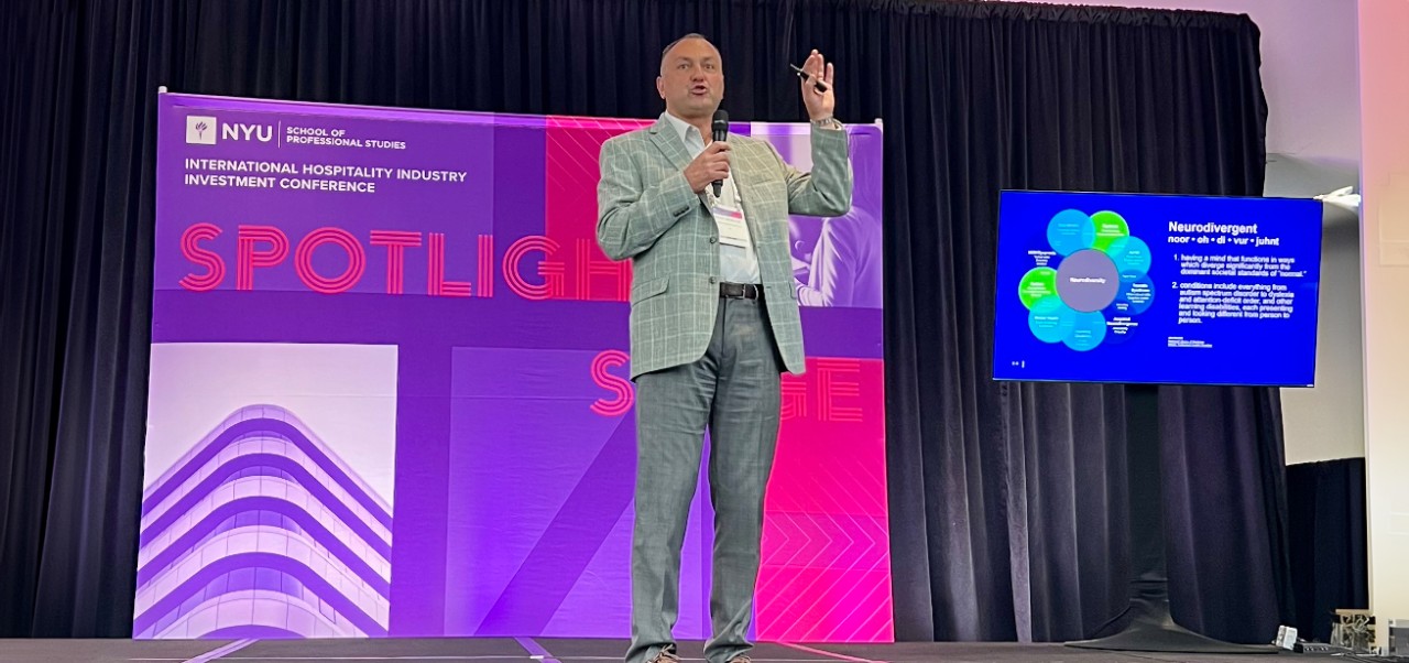 Recep Karaburun speaks with a microphone and one hand raised on a stage with a purple and pink NYU International Hospitality Industry Investment Conference Spotlight Stage banner behind him.