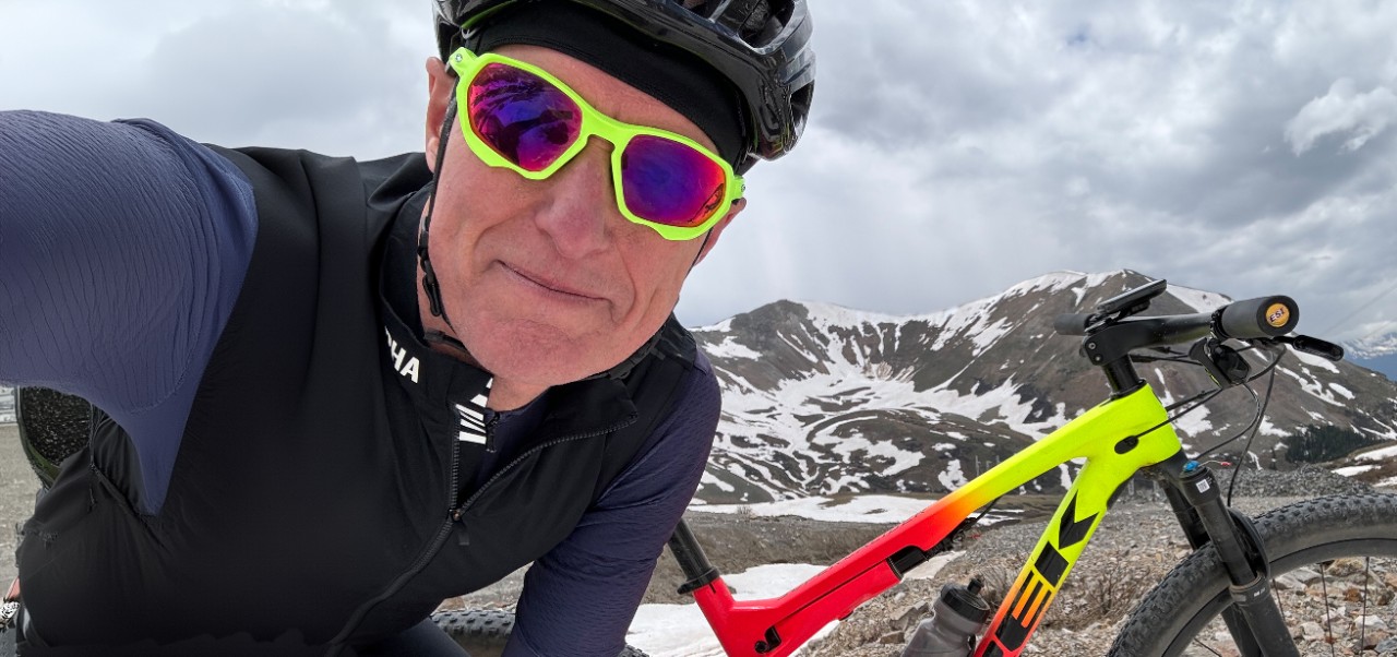 On a snowy mountaintop in Colorado, Scott Robinson wears green sunglasses and a helmet while taking a selfie with his red mountain bike behind him