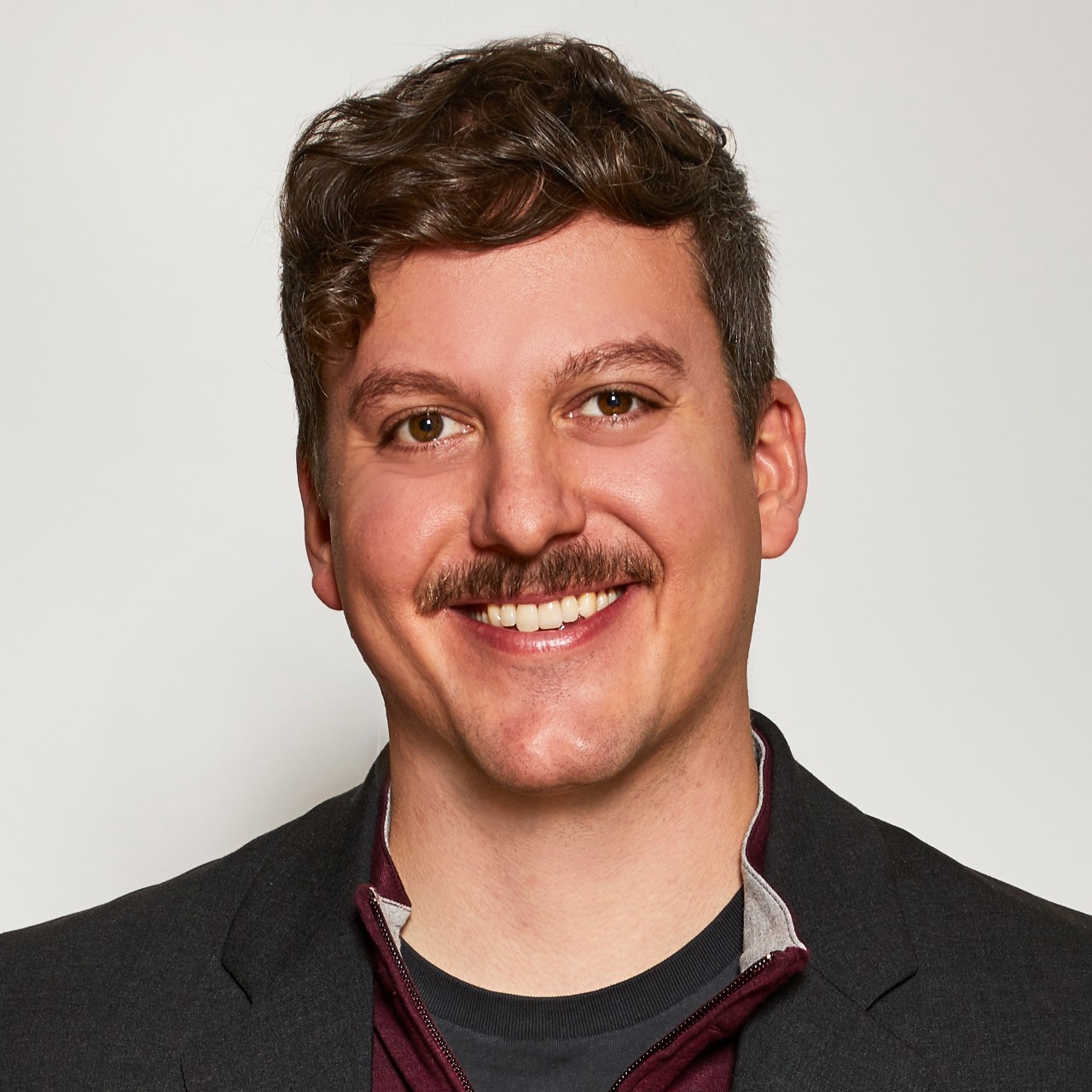 Simon Mullin smiles for a headshot in front of a solid white background while wearing a black blazer and a red half-zip shirt