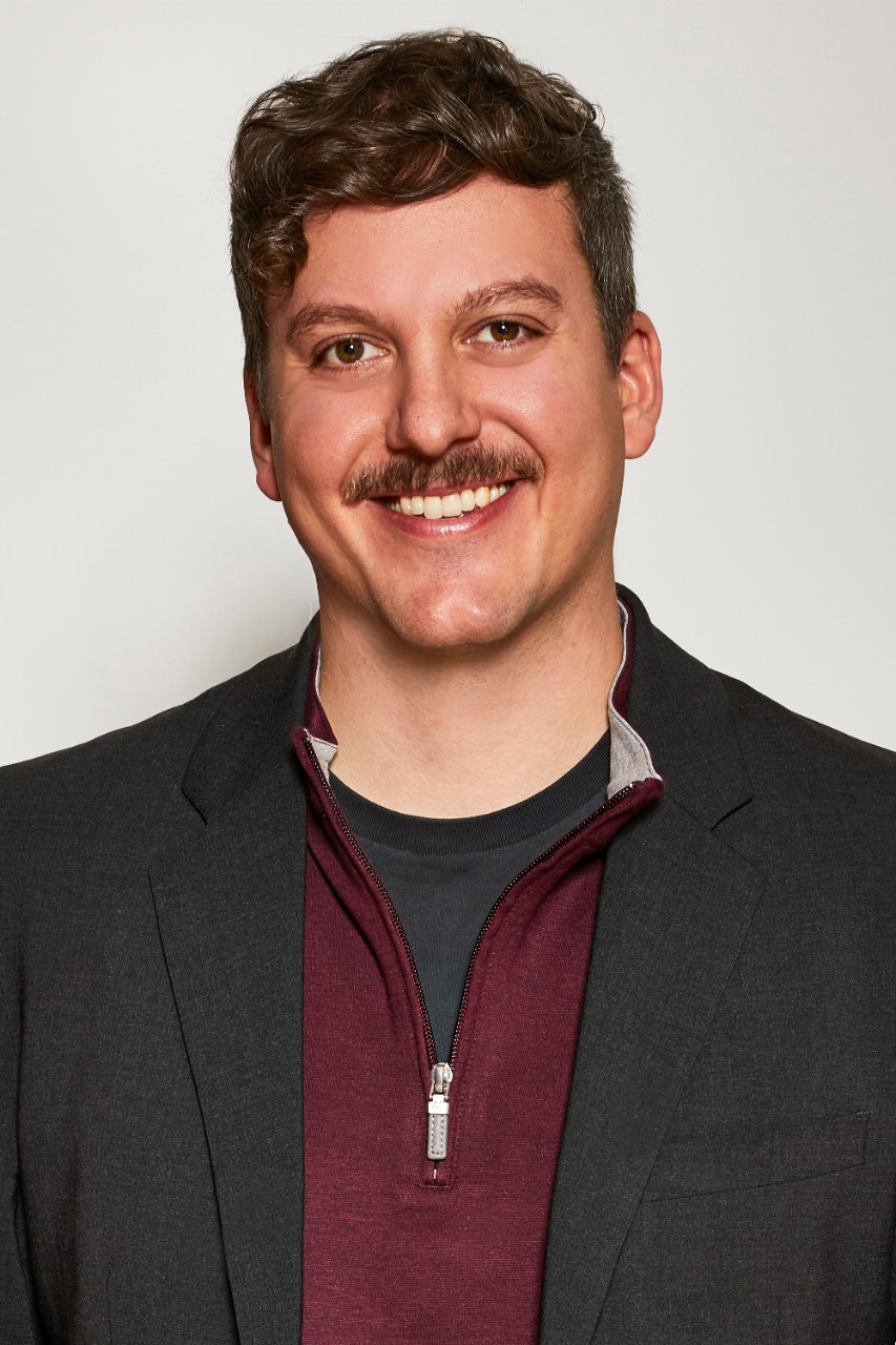 Simon Mullin smiles for a headshot in front of a solid white background while wearing a black blazer and a red half-zip shirt