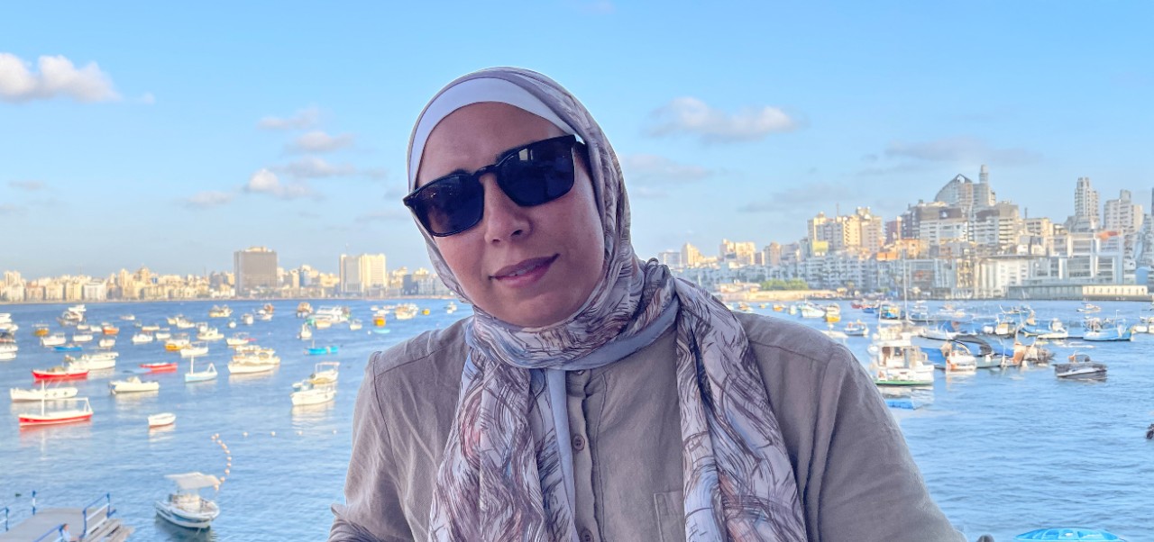  Yasmin Alkashef, Adjunct Instructor, Center for Publishing, Writing, and Media at NYU SPS, poses for a photo in front of a busy, gleaming, urban harbor full of watercraft.