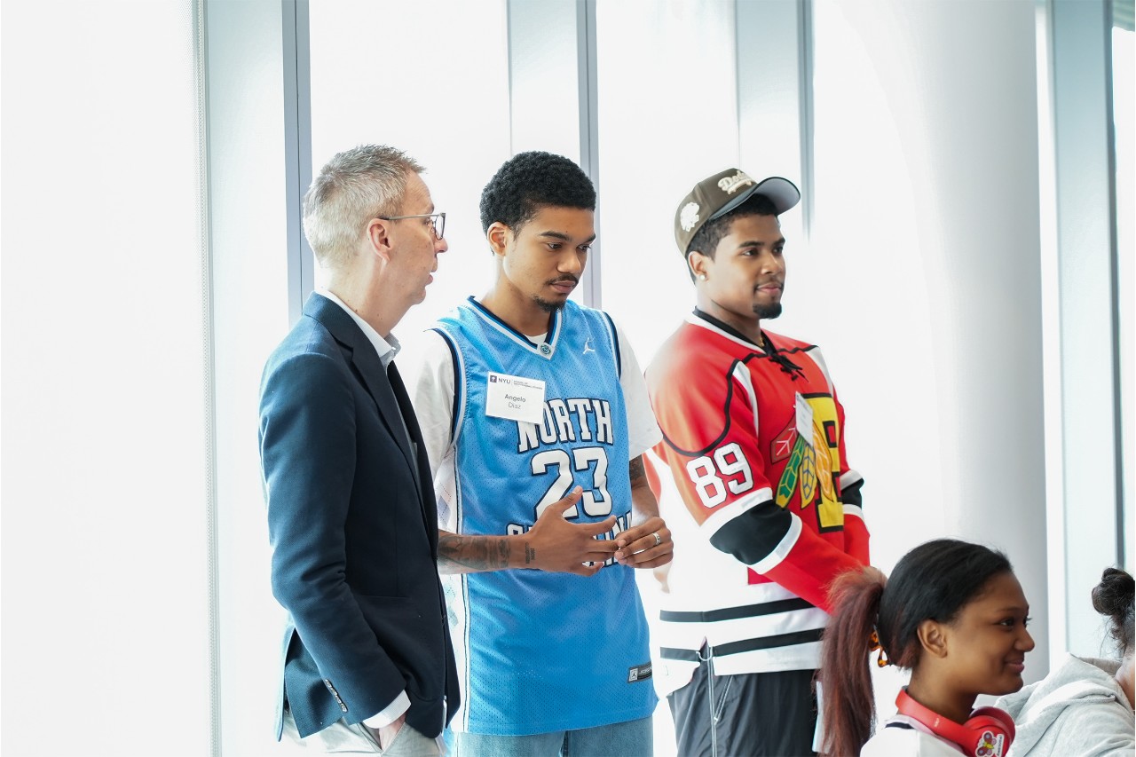Three men standing by a window, two wearing sports jerseys and one in a blazer.