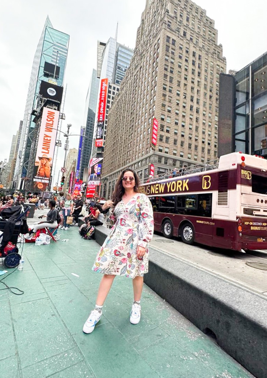A woman in a floral dress and sneakers smiles while standing in Times Square, New York City.