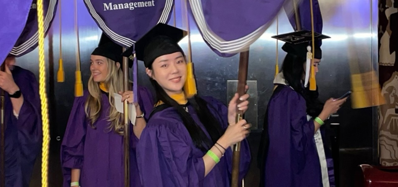Ji Yeon Baik, Center for Publishing and Liberal Arts at NYU SPS student holds an SPS flag while leading a procession of graduates at graduation.