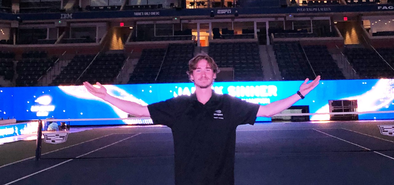 Peter Dicce, student at the Preston Robert Tisch Institute for Global Sport at NYU SPS, poses for a photo while standing in an empty, indoor tennis court at night.