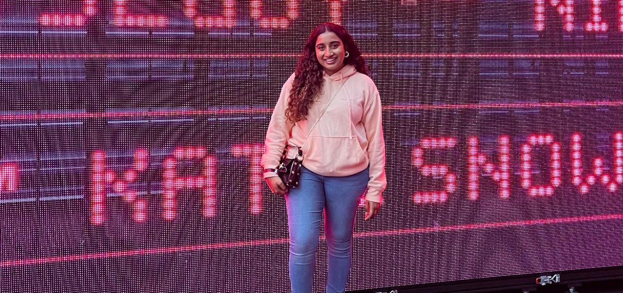 Stuti Daga, student at the Robert Tisch Institute for Global Sport at NYU SPS, smiles for the camera while standing in front of a massive digital display conatining pink letters.