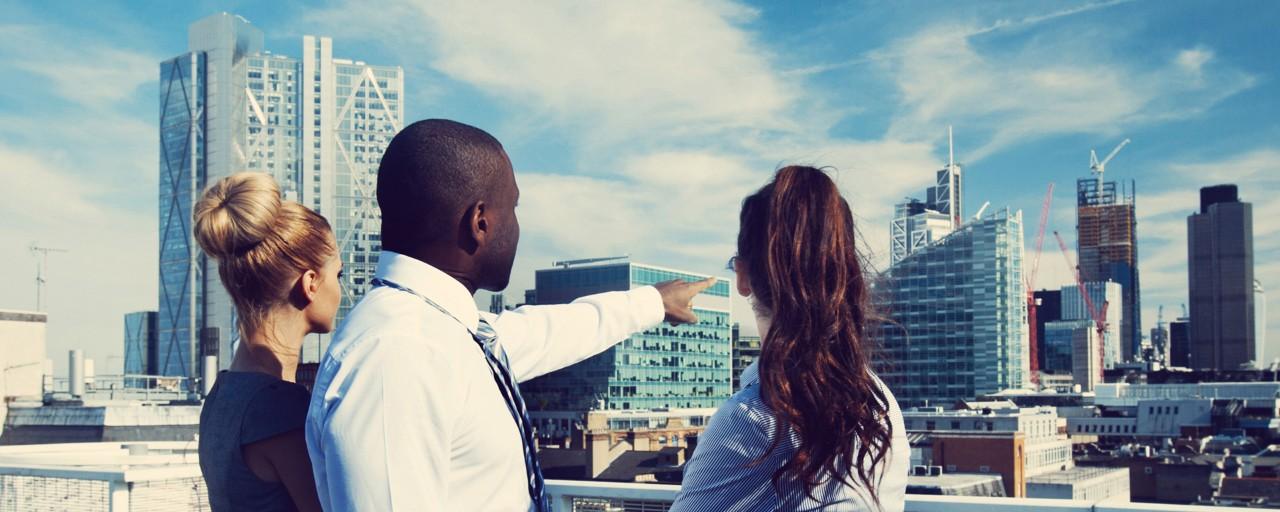 On a NYC rooftop, two women stand on either side of a man who is pointing toward skyscrapers in the distance