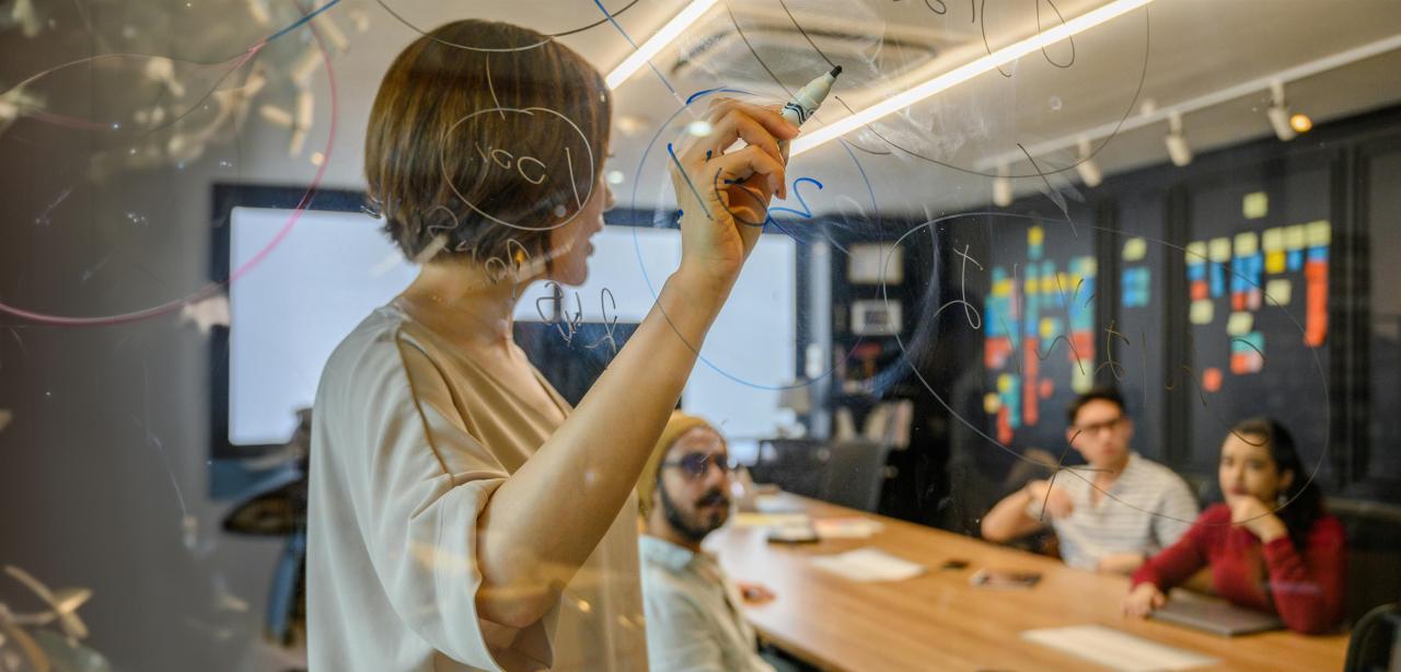 A woman draws on a transparent white board while demonstrating a business concept to colleagues seated at a table in the background.