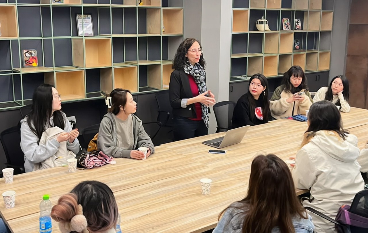 A group meeting in a modern conference room with several people seated around a large table and one woman standing and speaking.