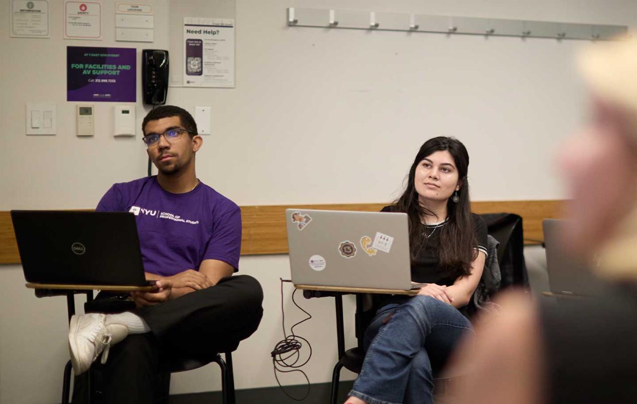 A classroom scene with two students seated with laptops, attentively listening.
