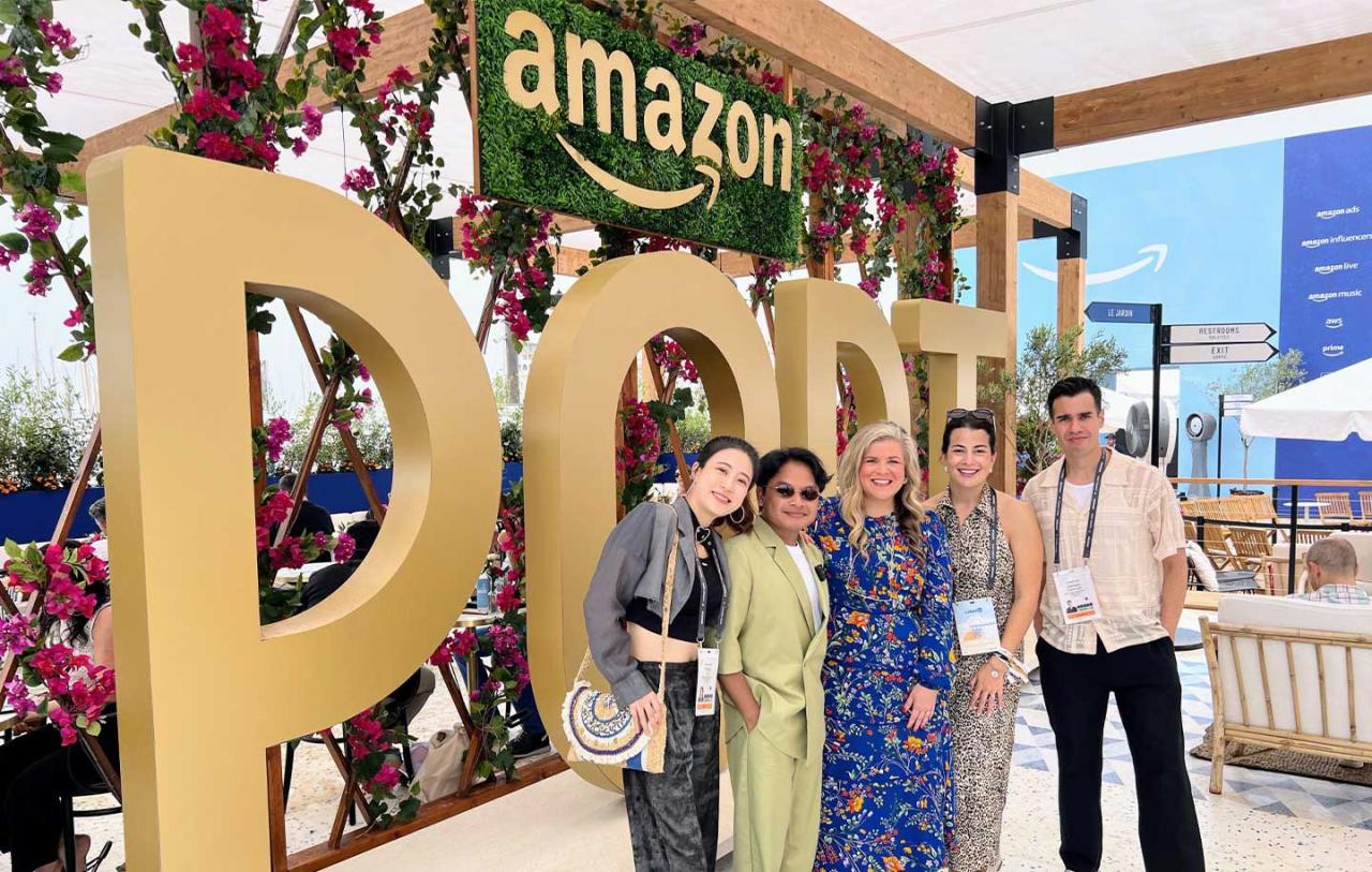 A group of five people stands smiling in front of a large gold Amazon sign adorned with pink flowers.