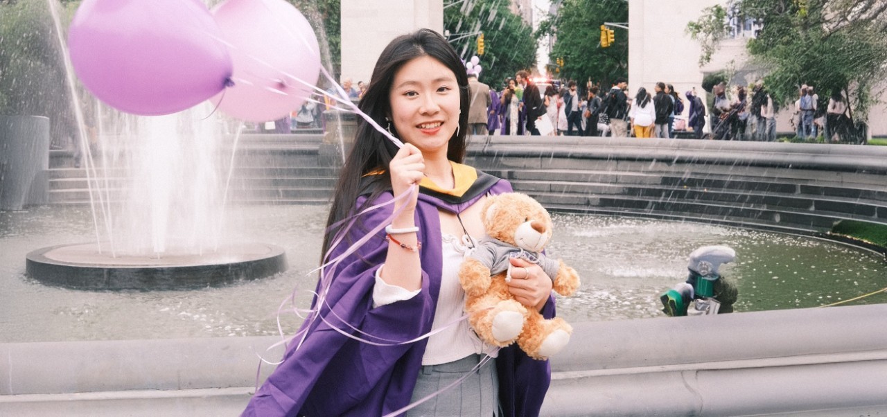 Angel Ma, Division of Programs in Business at NYU SPS alumnus, poses for a photo on graduation day in Washington Square Park while holding a teddy bear and balloons.