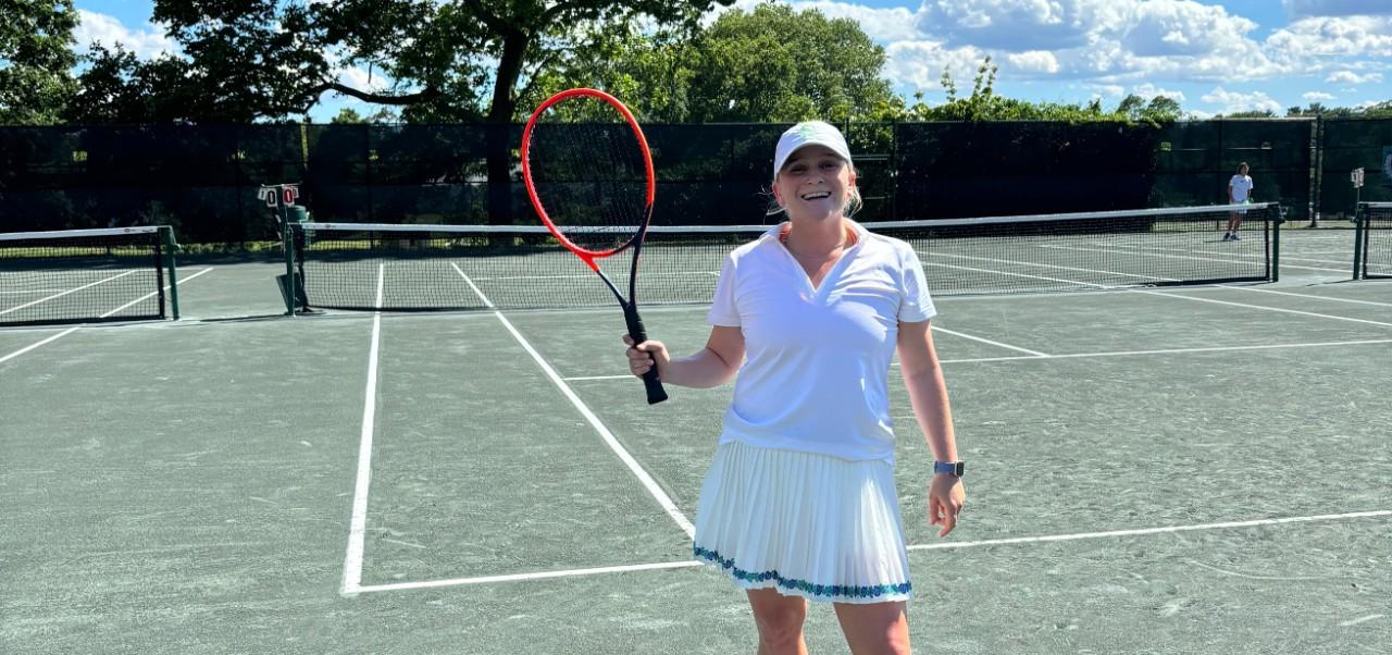 Caroline Kasper, NYU SPS Preston Robert Tisch Institute for Global Sport alumnus, takes a break from playing tennis to smile for the camera while holding her racket.