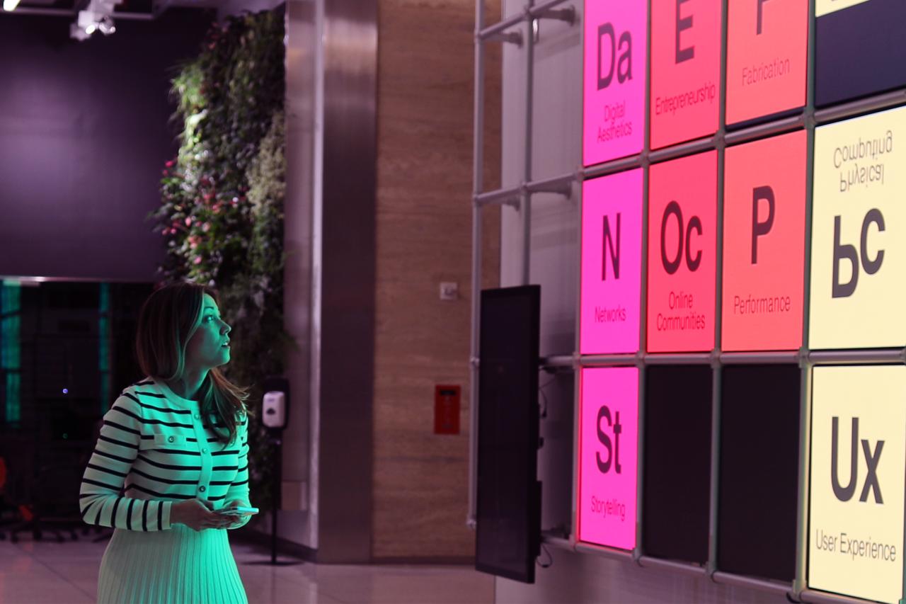 Faculty member Piper Henriques looks at a colorful, illuminated wall display with various abbreviated text panels in a modern interior featuring a green plant wall.
