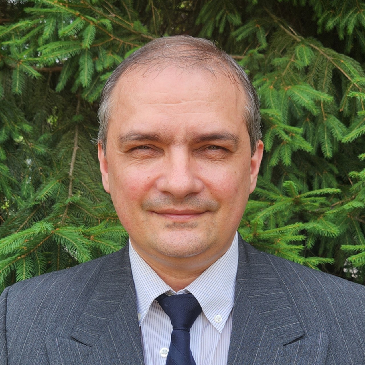 Bohdan Hawryluk softly smiles while wearing a suit and standing in front of a pine tree