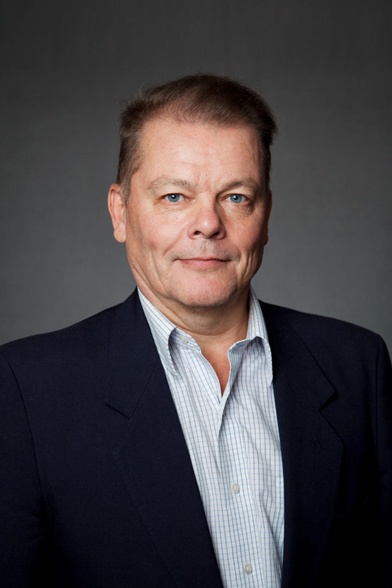 Jukka Laitamaki poses for a headshot in front of a grey background while wearing a black suit jacket and a blue grid-patterned shirt
