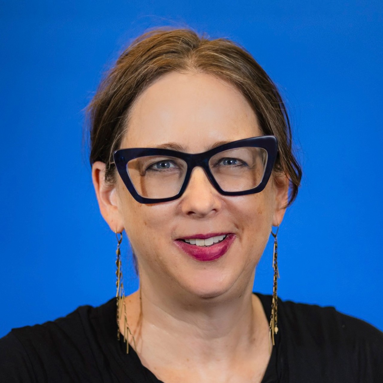 Julie Torres Moskovitz smiles for a headshot while wearing a black shirt in front of a solid blue background