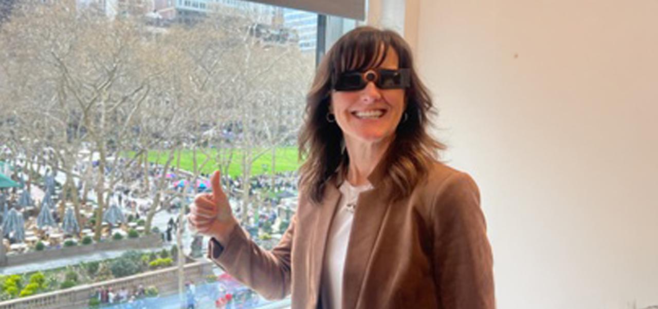 Kathleen Rennie, Clinical Assistant Professor of Marketing and Public Relations, Division of Programs in Business at NYU SPS, gives a "thumbs up" to the camera while standing in front of a window overlooking a park.