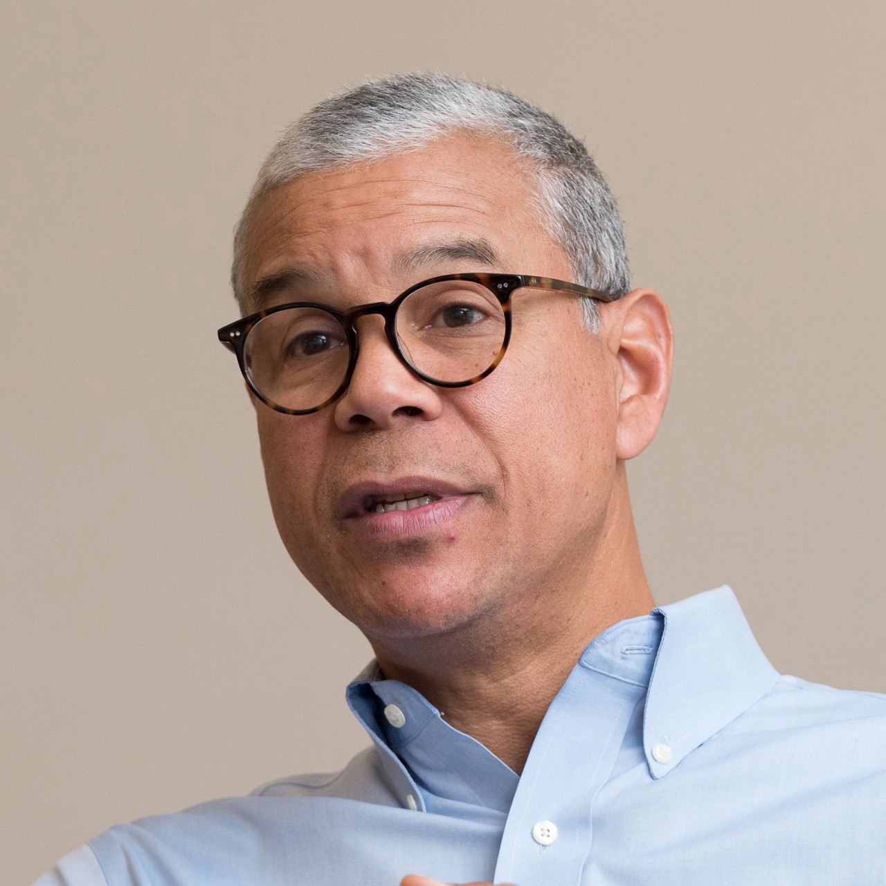 Michael Valentine folds his hands in front of him while wearing a light blue shirt in front of a beige background.