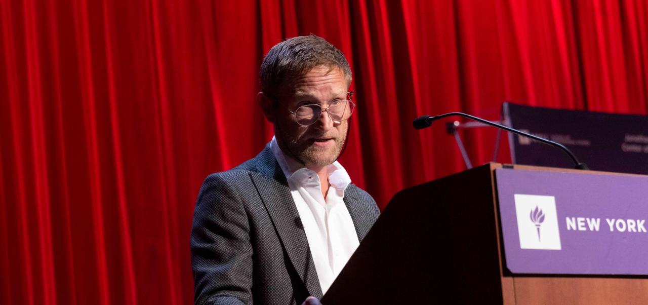 Nicolas Graf, Clinical Professor, Jonathan M. Tisch Center of Hospitality at NYU SPS addresses an audience from a lectern in front of a red curtain.