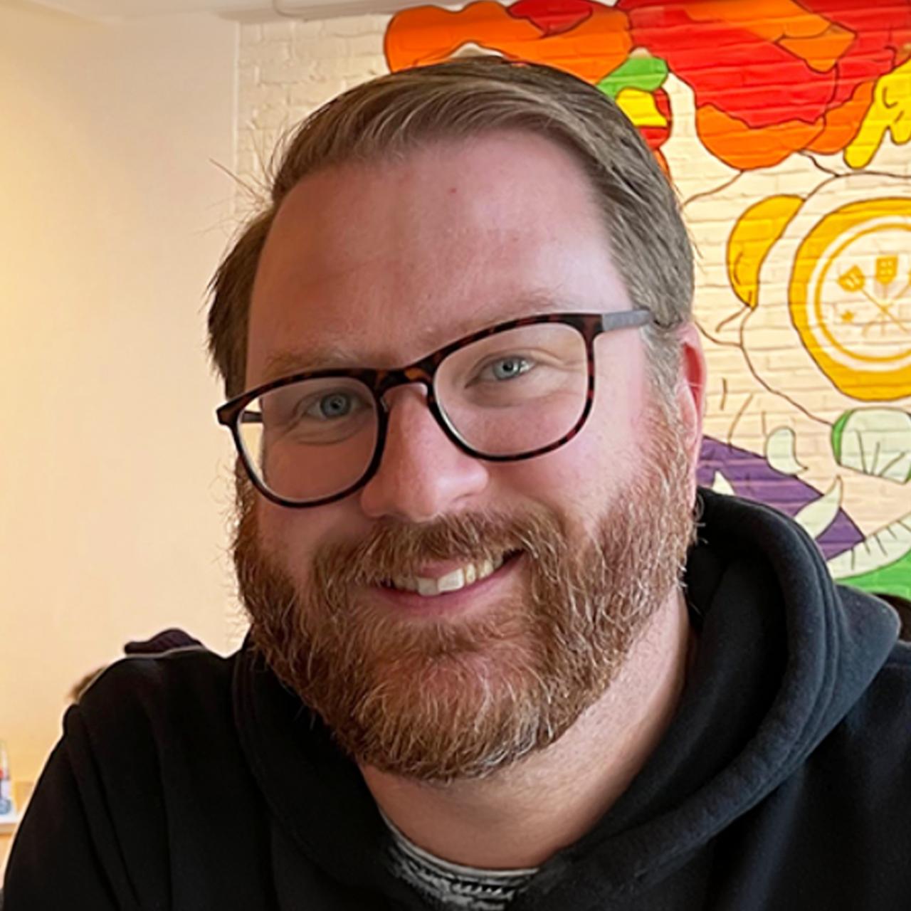 Patrick Gray smiles for a photo while wearing a black zippered jacket in front of a mural in the background