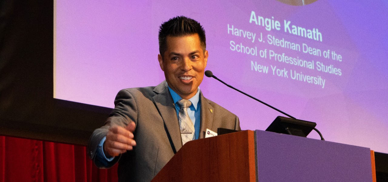 Raul Sanchez speaks into a microphone onstage behind a purple podium with a red curtain and a screen behind him at the NYU SPS Innovation Day event