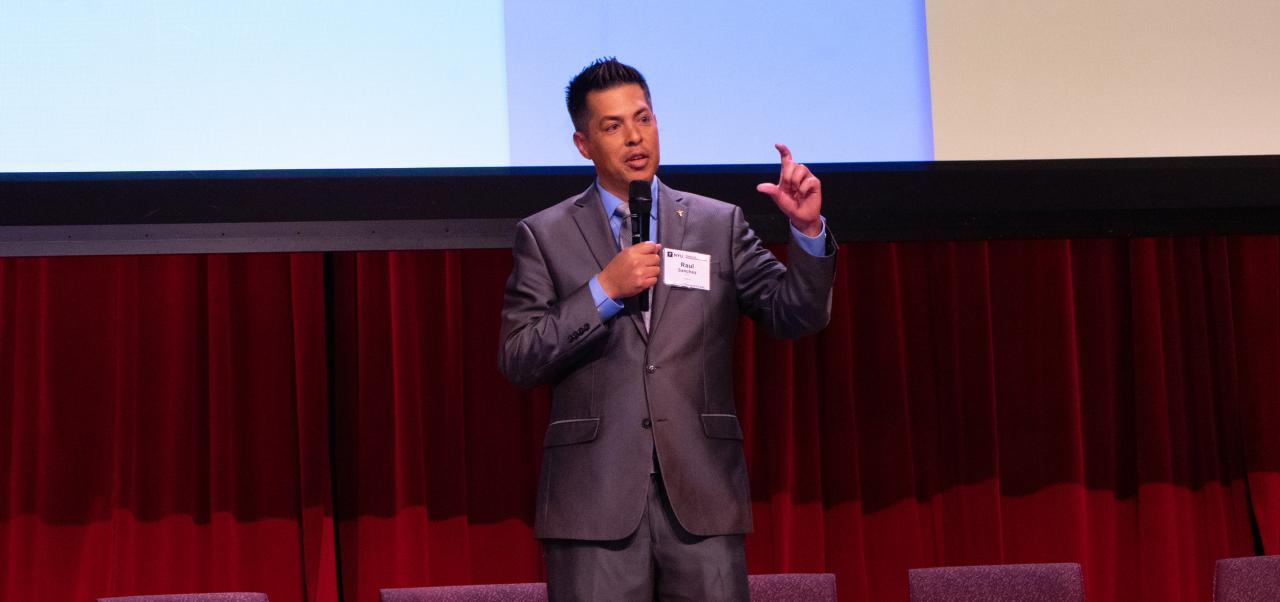 Raul Sanchez stands on a stage with a red curtain and a screen behind him at the NYU SPS Innovation Day event