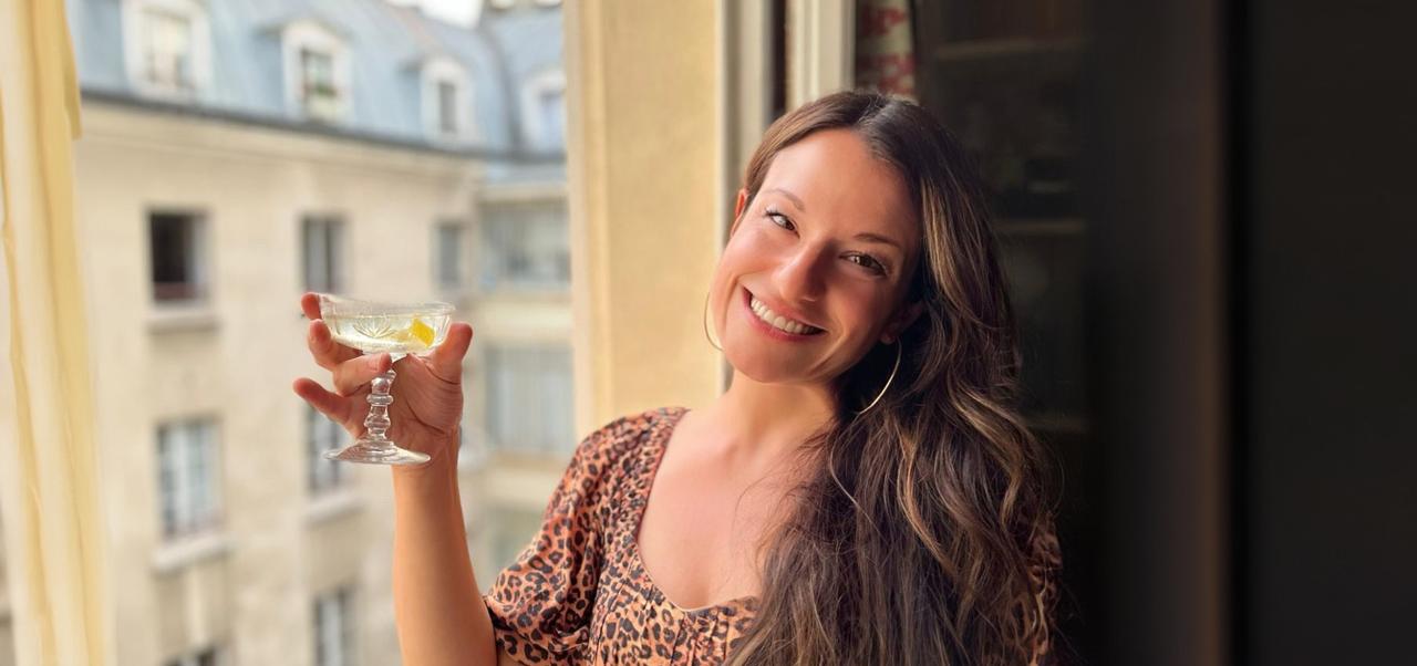 Tracy Olenick smiles next to a window with a classic Paris building in the background while holding a drink in her hand.