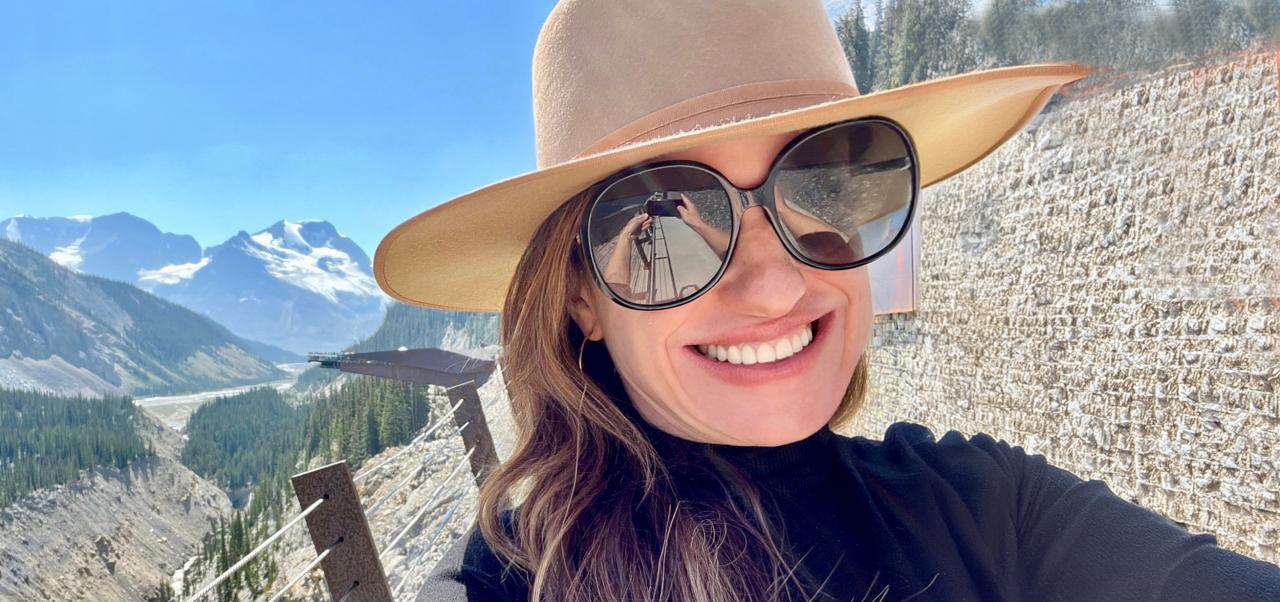With the Rocky Mountains behind her, Tracy Olenick wears sunglasses and a hat while taking a selfie.
