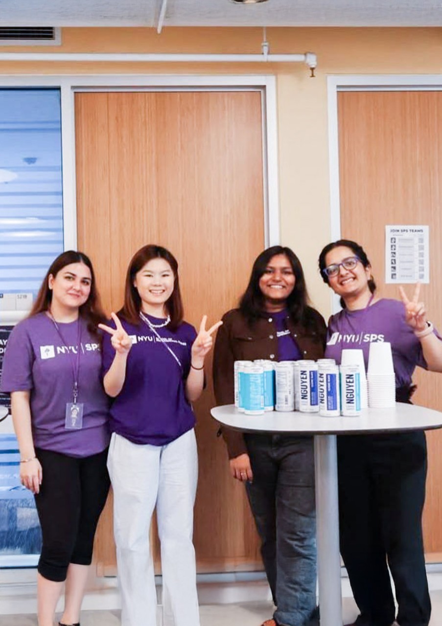 Four people in purple NYU shirts smile and pose with peace signs indoors.