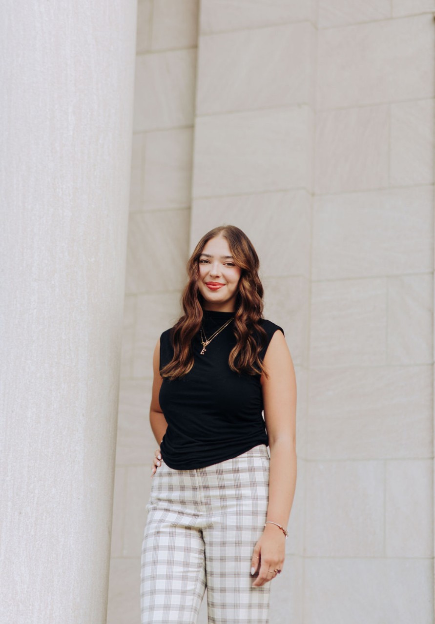 Ellie Sabby, wearing a sleeveless top and tartan slacks, poses  against a marble wall.
