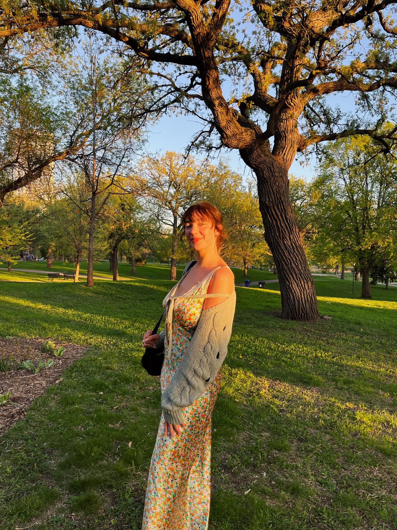Ellie Sabby, wearing a dress and carrying a purse, poses for a photo in a park.