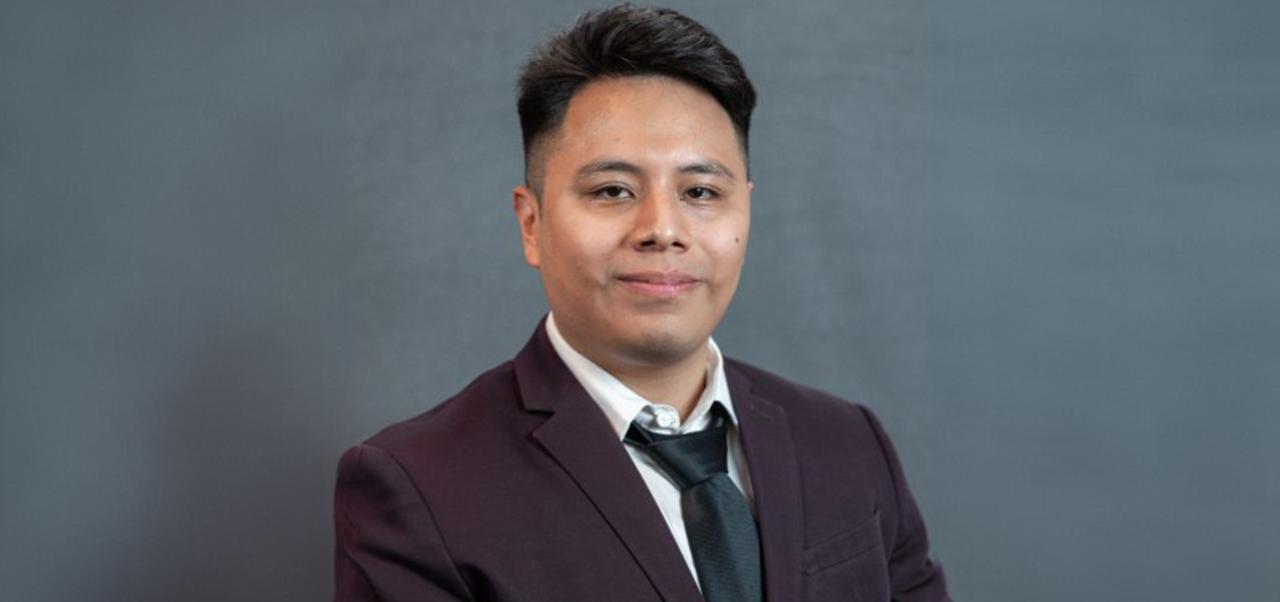 Jonathan Hernandez Jimenez smiles for a headshot wearing a dark grey jacket and tan shirt