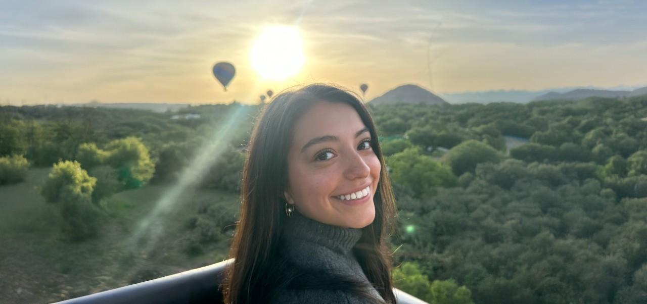 Nicole Landeta, Preston Robert Tisch Institute for Global Sport at NYU SPS alumnus, smiles for the camera in front of a dramatic sunset dotted by hot air balloons.