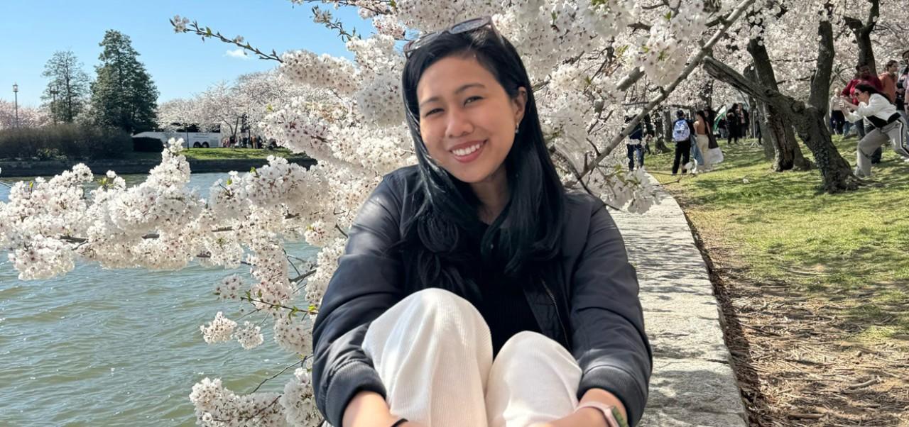 Nurul Syuhaidah Binte Senin, Jonathan M. Tisch Center of Hospitality at NYU SPS student, smiles for the camera while sitting at a pond amongst blooming cherry blossoms.